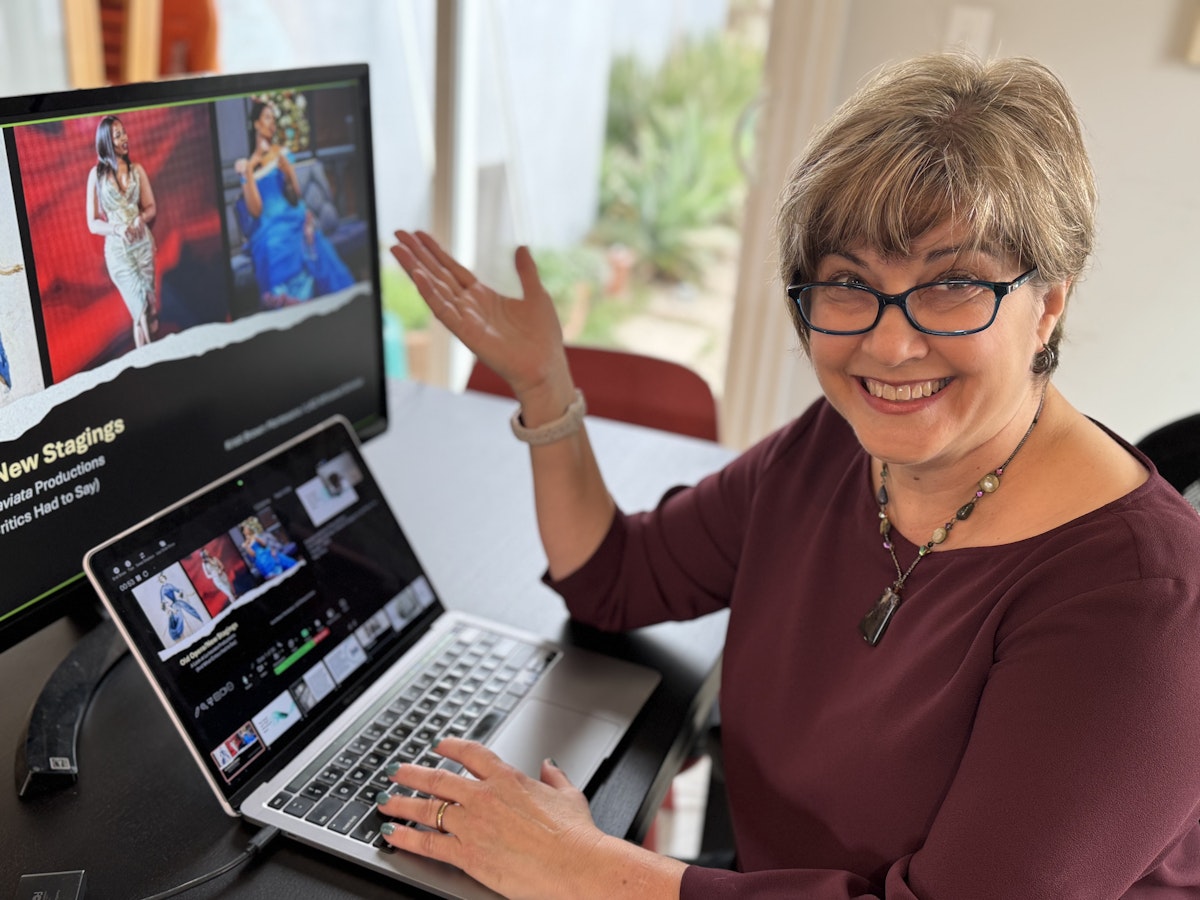 Woman with short hair, glasses, and a maroon shirt sitting in front of a laptop and desktop monitor showing the LA Opera website. She is smiling and gesturing towards the screens.